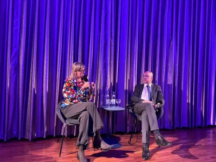 Krušnica, razgovor na događaju u amfiteatru s plavičastim zavjesama, žena i muškarac na panel diskusiji.