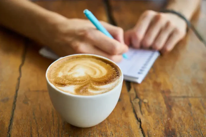 Mliječna kafa u šolji na drvenom stolu, osoba piše u notesu, kafa na temu krušnica, Bosna i Hercegovina.