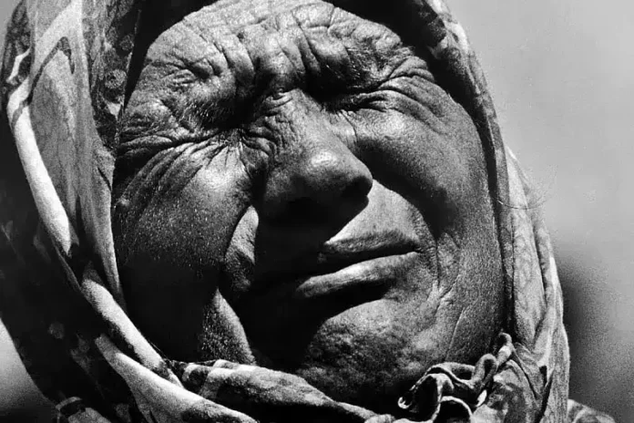 Muškarac s ocvalim licem, blago nasmiješen, nosi tradicionalnu odjeću i šešir, u crno-bijeloj fotografiji.