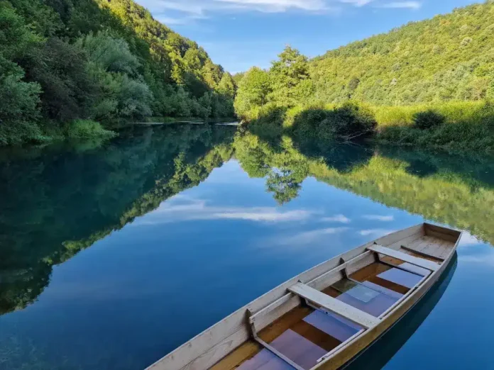 Mirna rijeka uz obalu sa zelenim brdima, plaža i drveni čamac u prirodnom okruženju.
