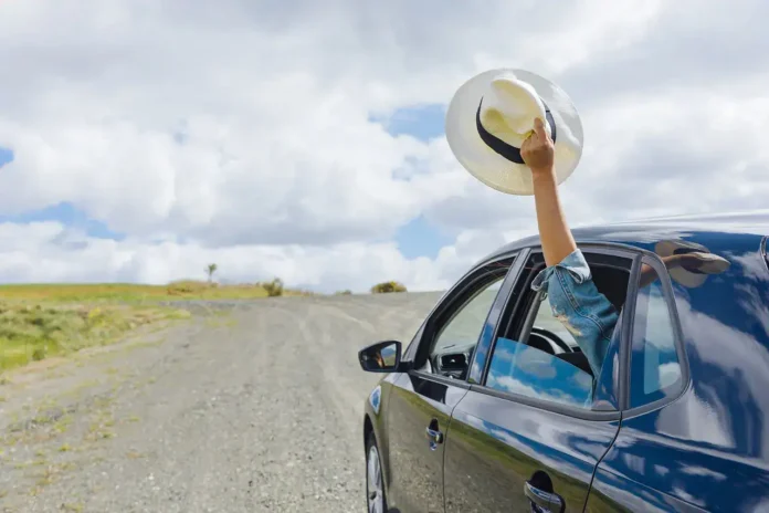 Osoba s šeširkom na ruci izlazi iz automobila na ruralnom putu uz prirodu.