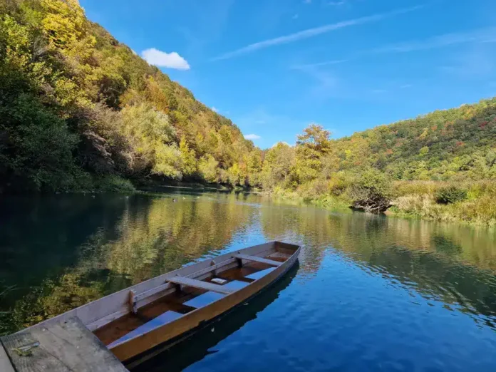 Krušnica rijeka u prirodi, pored drvene čamce, jesenja zelena i plavi nebeski odsjaj.