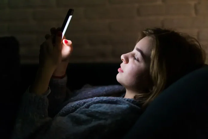 Lijevo, mlada žena koristi mobilni telefon u mraku, osvjetljenje iz ekrana ističe njen lice.