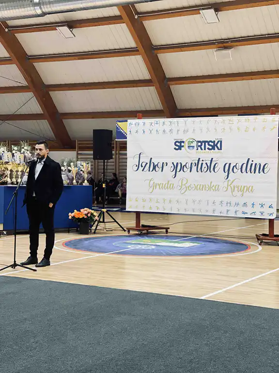 Muškarac u odijelu drži mikrofon na sportskom događaju u dvorani.