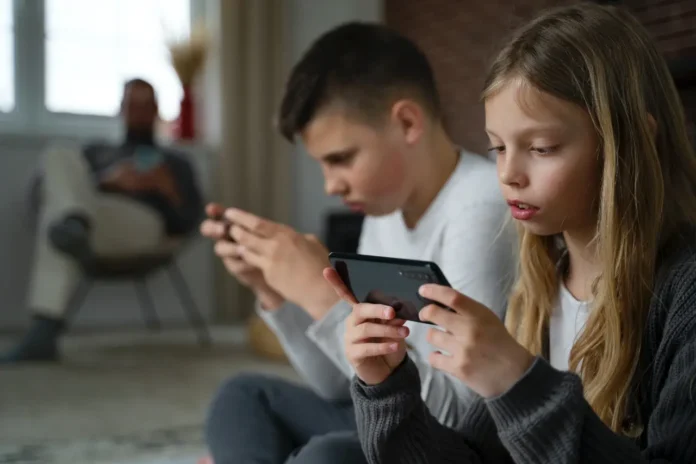 Djeca koriste mobilne uređaje dok sjede u dnevnoj sobi, odražavajući modernu tehnologiju i svakodnev.