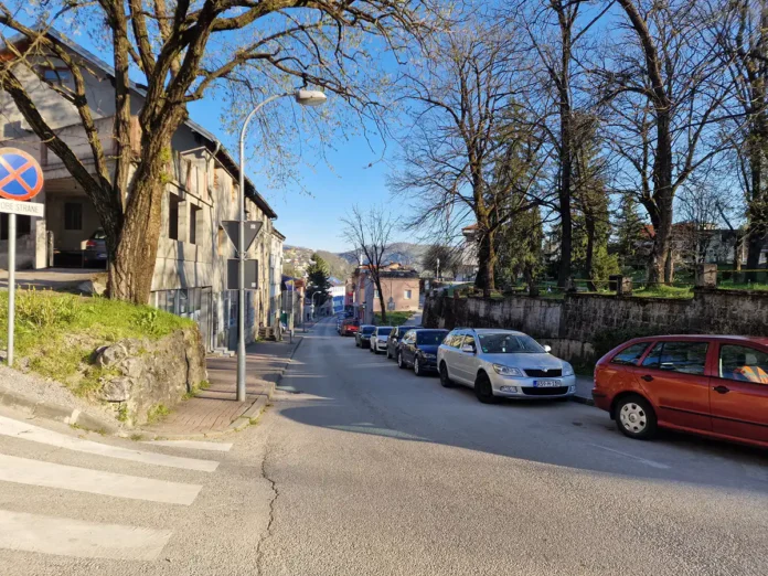 Ulica u Bosanskoj Krupi s parkiranim vozilima i drvećem u pozadini.