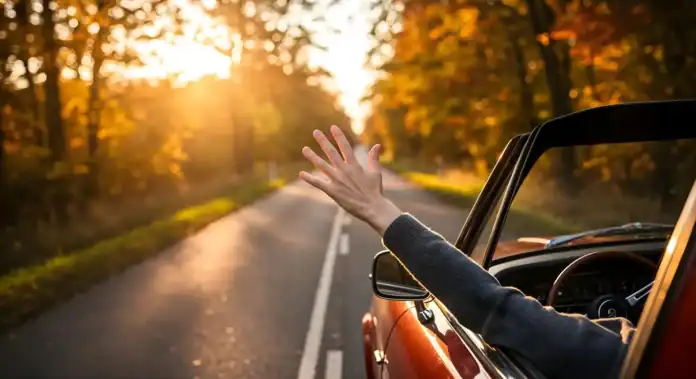 Vozač maše s prozora automobila na opuštenoj cesti u jesenjem okruženju.