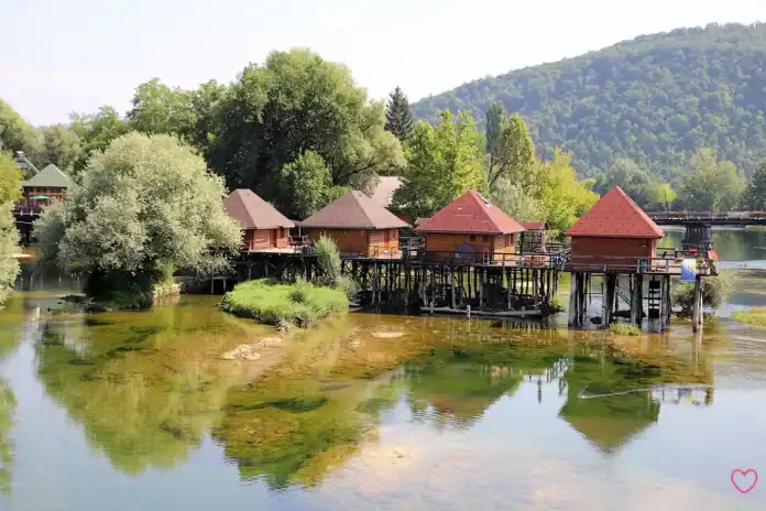 Krušnica rijeka s drvenim kućama na obali i zelenim brdima u pozadini.